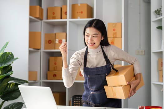 A joyful young female entrepreneur celebrates success in her workspace, holding packages while working on her laptop.