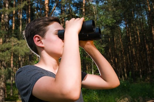 Side head-and-shoulder view of a 13-year-old male teenager looking through binoculars in the woods