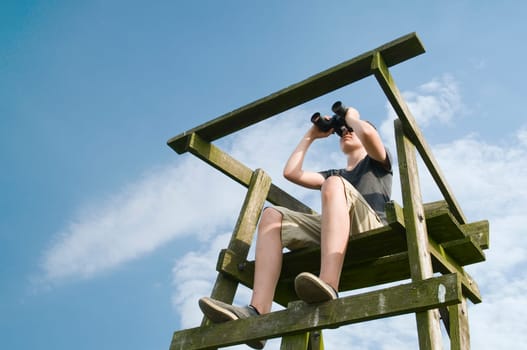 Full-body view of a 13-year-old male teenager sitting on an old high-rise in the summer and looking through binoculars in front of blue skies with individual white clouds