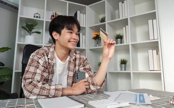 A young man smiles while holding a credit card, indicating successful financial management in his home office, representing positive finance habits.