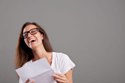 brunette girl with glasses laughing about news she received on a white document on a neutral gray faded background