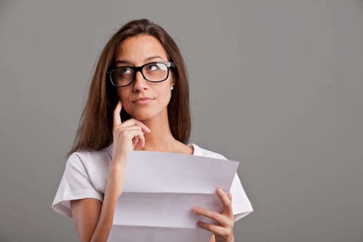 brunette girl with glasses thinking about news she received on a white document on a neutral gray faded background