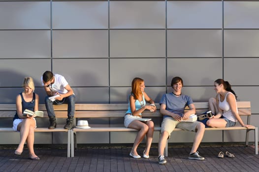 College students sitting on bench by modern wall outside campus