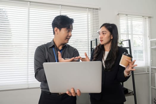 Two business professionals conversing and reviewing information on a laptop while one holds a coffee cup in a collaborative office setting.