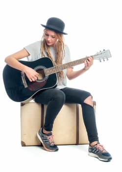Blonde girl with hat sits on a suitcase and plays guitar
