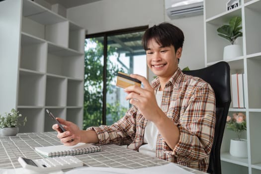 A joyful young man holds a credit card and smartphone, celebrating financial success in his home office, representing positive finance management.