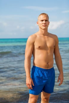 Young man in blue swimming trunks stands on the beach in the blue sea on holiday