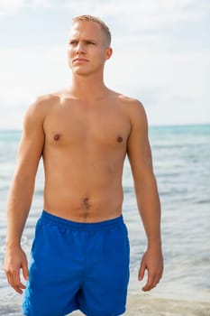 Young man in blue swimming trunks stands on the beach in the blue sea on holiday