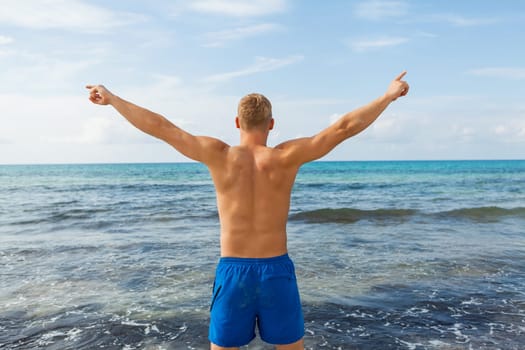 Young man in blue swimming trunks stands on the beach in the blue sea on holiday