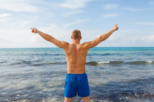 Young man in blue swimming trunks stands on the beach in the blue sea on holiday