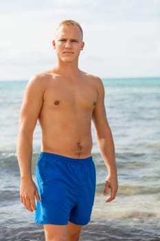 Young man in blue swimming trunks stands on the beach in the blue sea on holiday