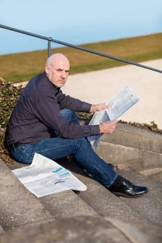 Middle-aged man,senior sitting with a newspaper on a staircase in spring outdoors