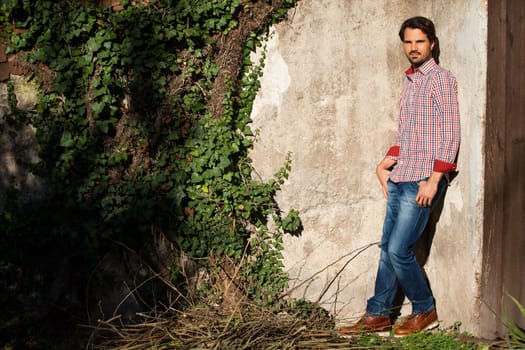 Attractive young man with a plaid shirt and jeans leaning against a wall outdoors in summer