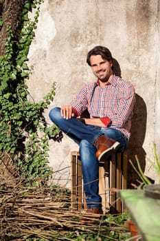 Young man with a plaid shirt and jeans with dark hair and beard outdoors under an efeuranke in daylight