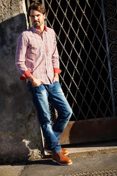 Attractive young man with a plaid shirt and jeans leaning against a wall outdoors in summer