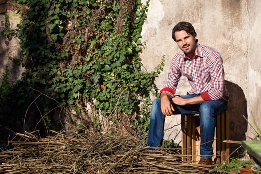 Young man with a plaid shirt and jeans with dark hair and beard outdoors under an efeuranke in daylight