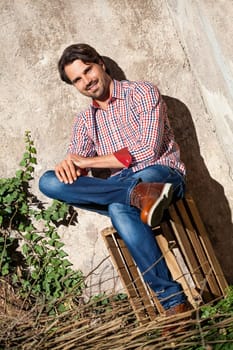 Young man with a plaid shirt and jeans with dark hair and beard outdoors under an efeuranke in daylight
