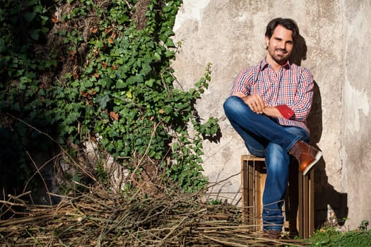 Young man with a plaid shirt and jeans with dark hair and beard outdoors under an efeuranke in daylight