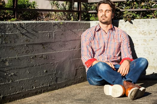 Young man with a checked shirt sits on a wall in the sun and relaxes