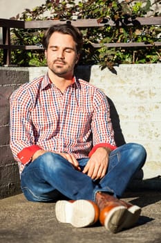 Young man with a checked shirt sits on a wall in the sun and relaxes