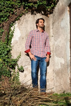 Young man with a plaid shirt and jeans with dark hair and beard outdoors under an efeuranke in daylight