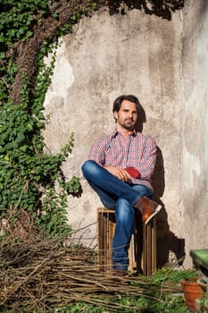 Young man with a plaid shirt and jeans with dark hair and beard outdoors under an efeuranke in daylight