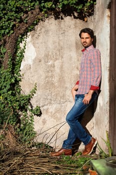 Young man with a plaid shirt and jeans with dark hair and beard outdoors under an efeuranke in daylight
