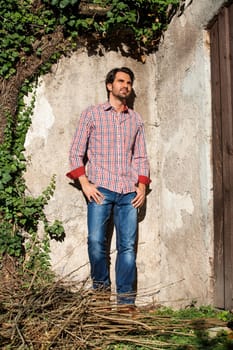 Young man with a plaid shirt and jeans with dark hair and beard outdoors under an efeuranke in daylight
