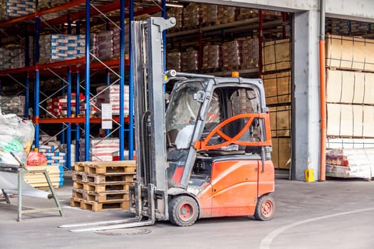 Small orange forklift in front of a warehouse with many pallets in daylight