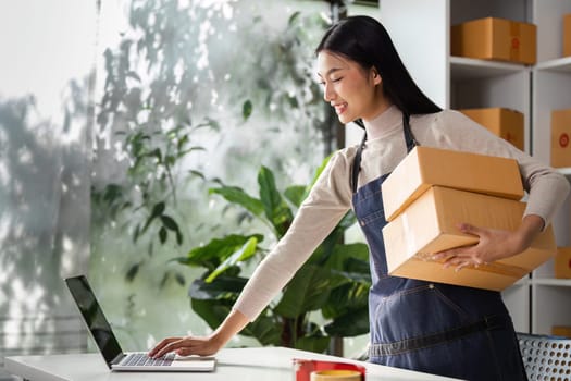 A young woman in an apron carries two boxes while working on a laptop in an organized workspace filled with greenery.