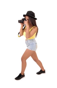 A young woman in shorts and a hat standing in the studio and taking.pictures with her camera, isolated over white background..
