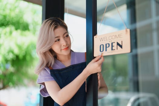 open a small business Portrait of happy Asian woman wearing apron holding welcome we are open sign at front of cafe