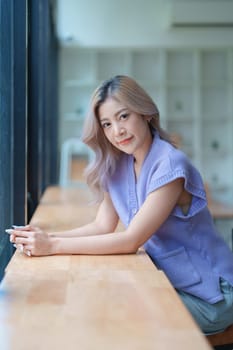 Asian young woman making notes in paper notebook sitting on cozy sofa on background of window. Pretty female writing notes in diary or making to do list relaxing at home