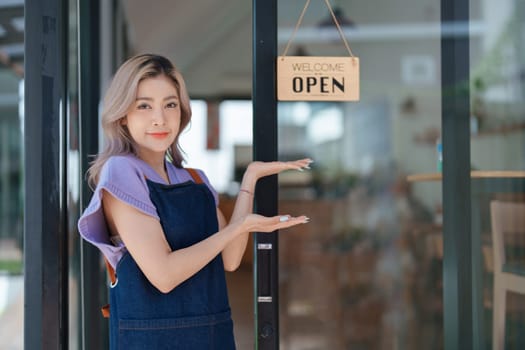 open a small business Portrait of happy Asian woman wearing apron holding welcome we are open sign at front of cafe