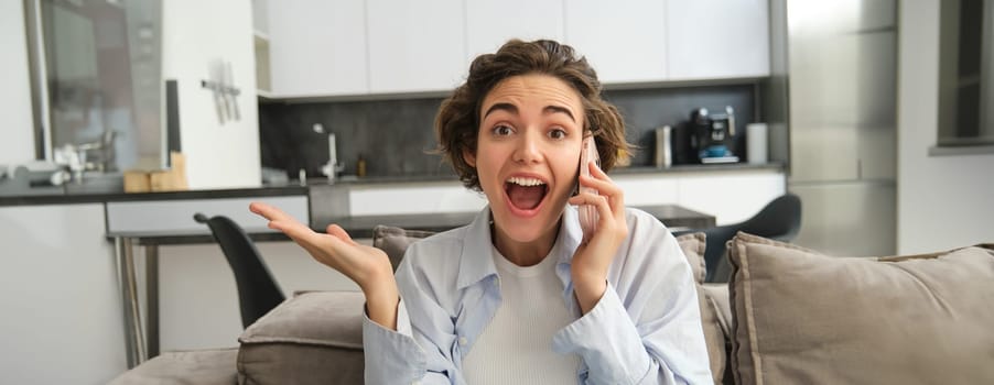 Close up portrait of amazed brunette woman, jumps on sofa from excitement, answers phone call, hear great news over the telephone, reacts surprised and happy.
