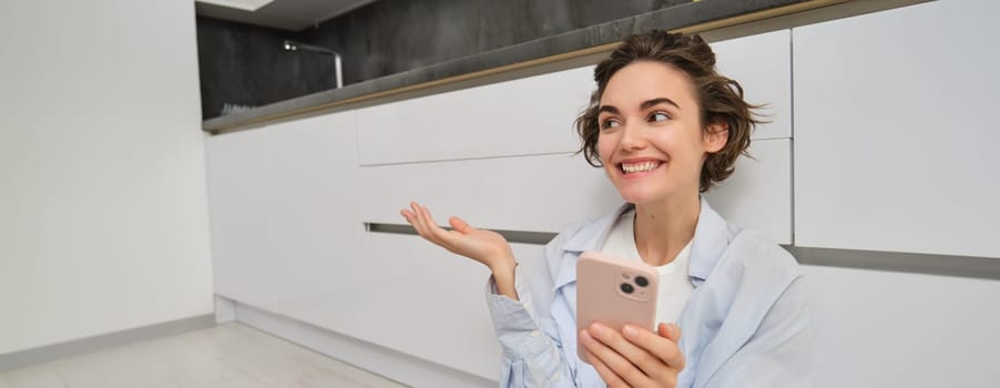 Portrait of happy young woman, shows her brand new kitchen, sits on floor, points at furniture and smiles, video chats on smartphone. Technology and lifestyle concept