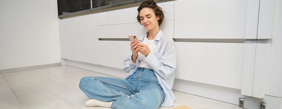 Portrait of girl with mobile phone, sits on floor at home, orders takeaway in app. Young woman with smartphone does online shopping from home, browsing websites.