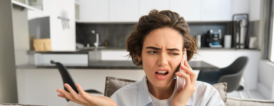 Image of woman with confused, annoyed face, talking on mobile phone while sitting at home on sofa, shrugging and looking bothered.