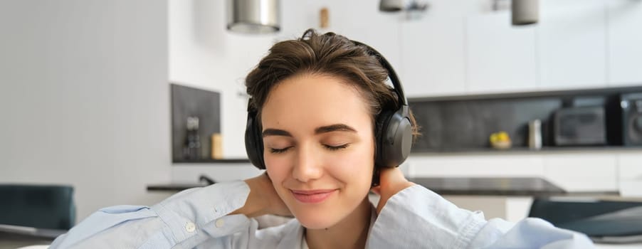 Close up portrait of girl with headphones, enjoys listen to music, close eyes and smiles form joy and delight.