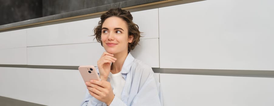 Portrait of girl with mobile phone, sits on floor at home, orders takeaway in app. Young woman with smartphone does online shopping from home, browsing websites.