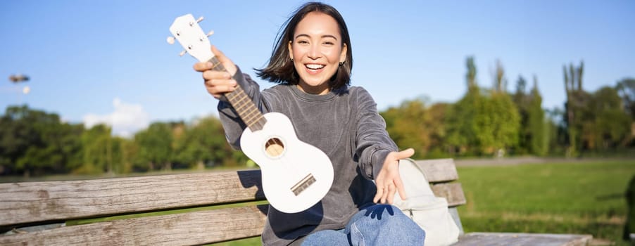 Happy asian girl shows ukulele, demonstrates her new musical instrument, sits on bench in park and plays music.