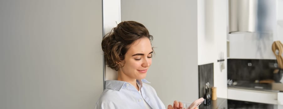 Portrait of smiling, beautiful woman, looking at smartphone, standing at home near kitchen, checking messages, chatting on mobile phone. Copy space