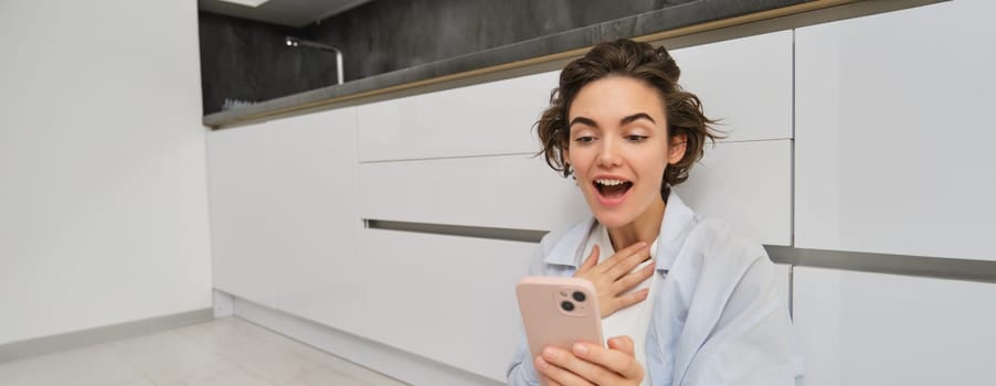 Portrait of girl looks at phone screen with amazed face. Woman reads news on smartphone with excitement, gasps, sits on floor at home.