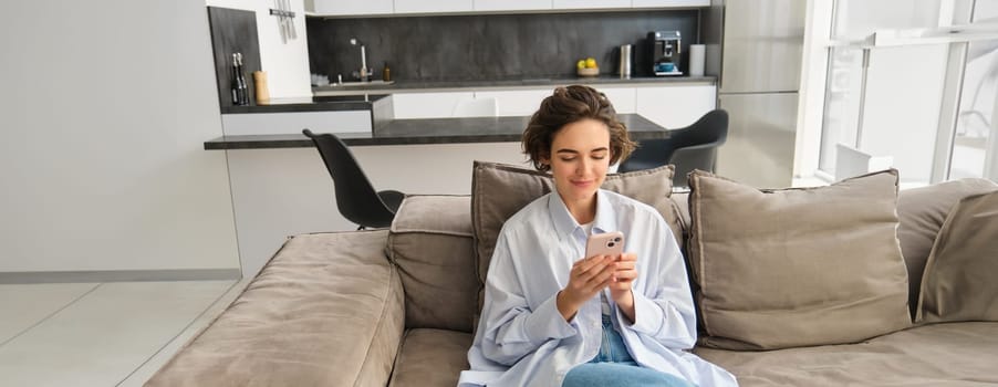 Portrait of brunette woman sits on sofa with smartphone, chats on phone, looks at her mobile app, checks messages on telephone, does online shopping from home.