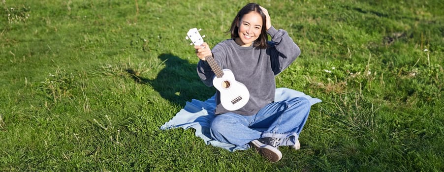 Excited asian girl sits in park with ukulele, plays instrument and feels happy upbeat, positive moments and people concept.