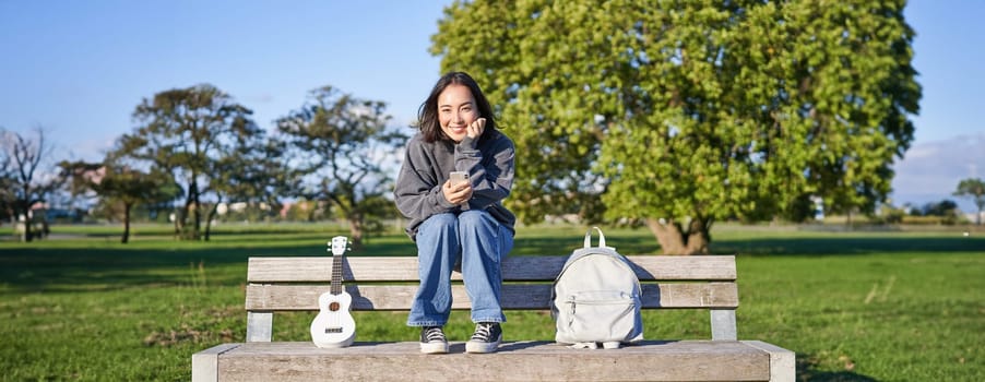 Beautiful brunette girl on bench in park, sitting with ukulele and backpack, holding smartphone, using mobile app.