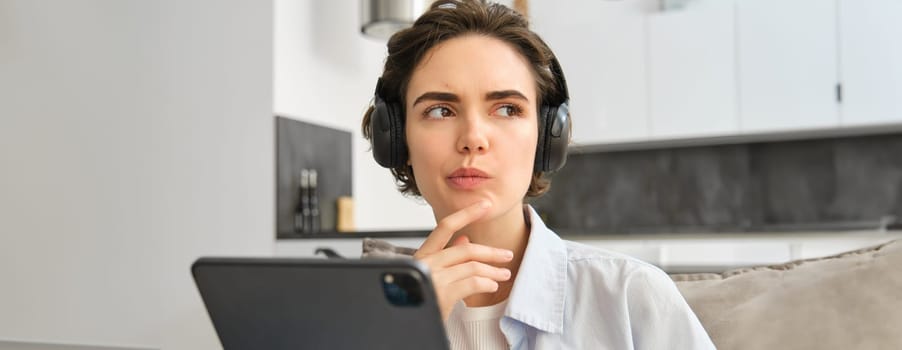 Portrait of woman studying, listening music in headphones, thinking while working on digital tablet, work remote from home. Copy space