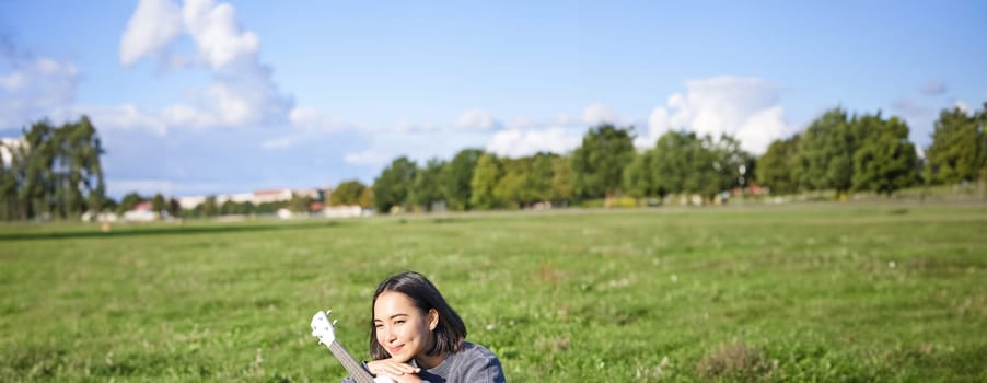 Romantic asian girl sitting with ukulele guitar in park and smiling, relaxing after university, enjoying day off on fresh air.