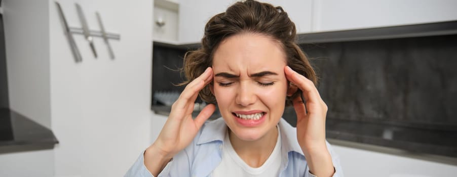 Portrait of woman with headache, sits in kitchen, touches head and grimaces, has painful migraine. Copy space