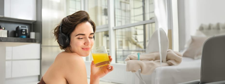 Smiling sportswoman, fitness girl does workout at home, wears headphones, listens music and drinks orange juice after training session.